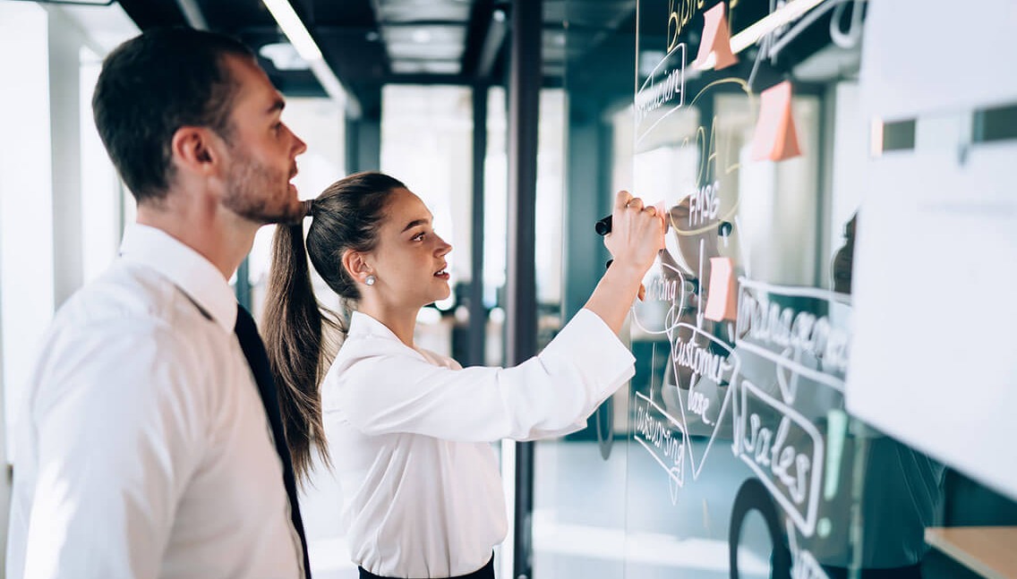 Side view of female drawing scheme of business strategy on glass board while male coworker observing process at modern workplace