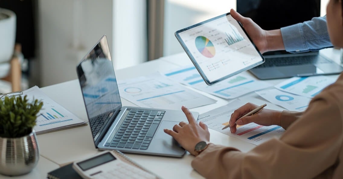 Two people looking at graphs and data on a laptop, tablet and paper