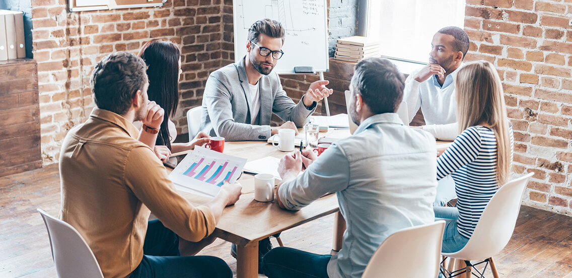 Group of six young people discuss something and gesturing while sitting at the table in office