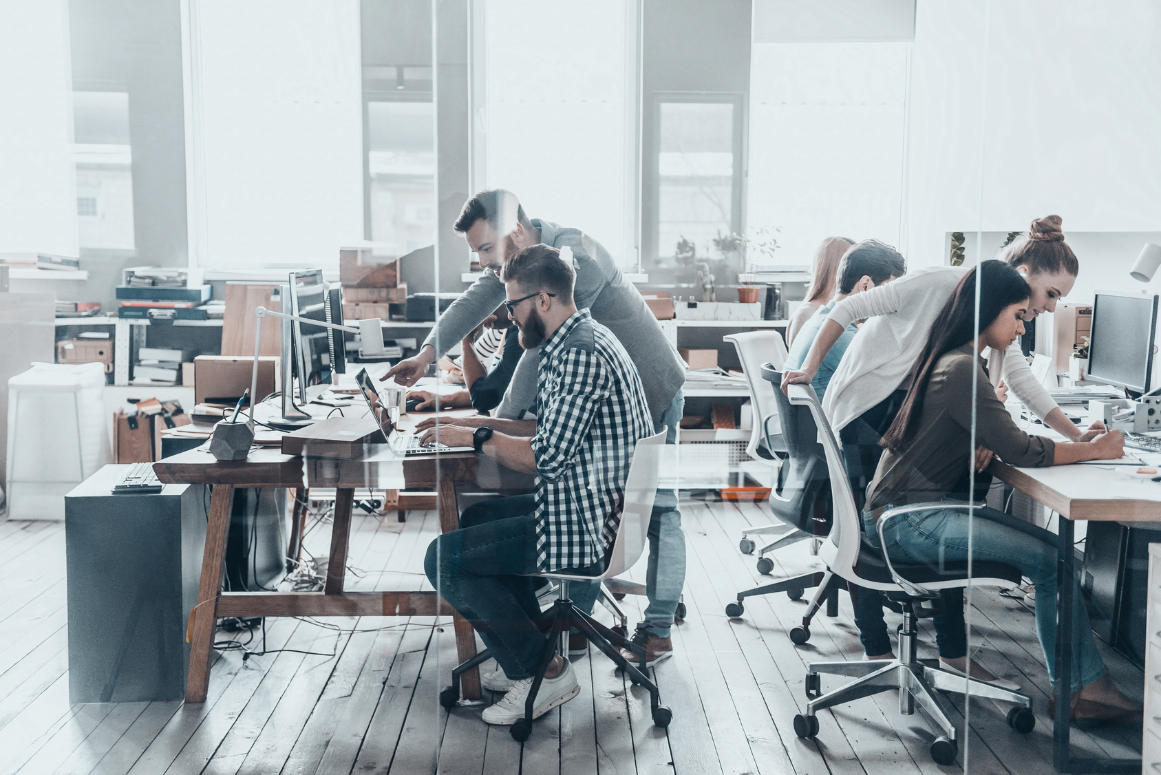 Group of young business people sitting at the office desk and discussing something while working together behind the glass wall in creative office.