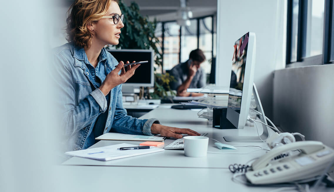 Young businesswoman talking on speaker phone and working on computer. Young woman sitting in front of monitor with smart phone.