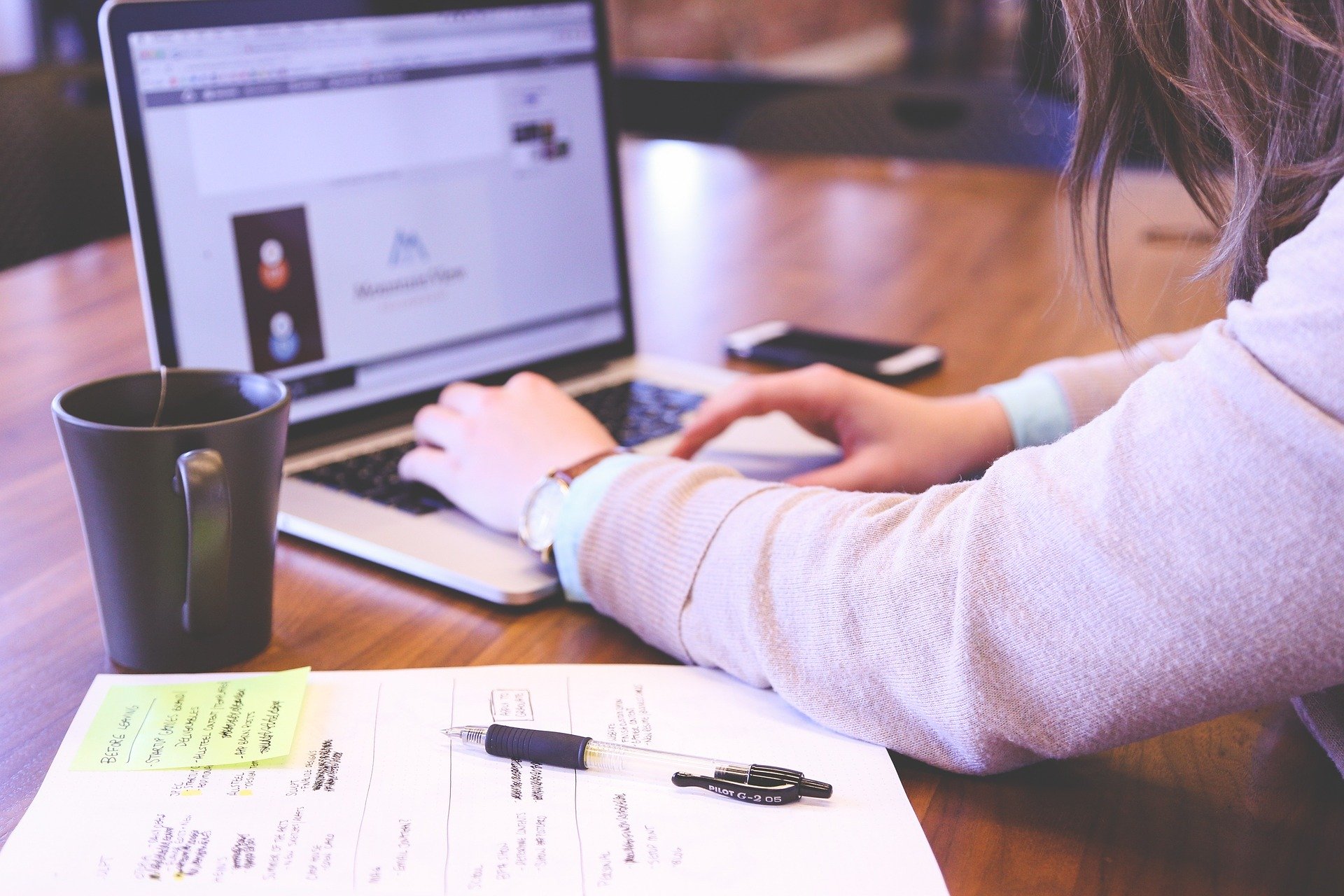 Working in a coffee shop at a laptop, with a pen and paper next to the laptop and a mug of tea