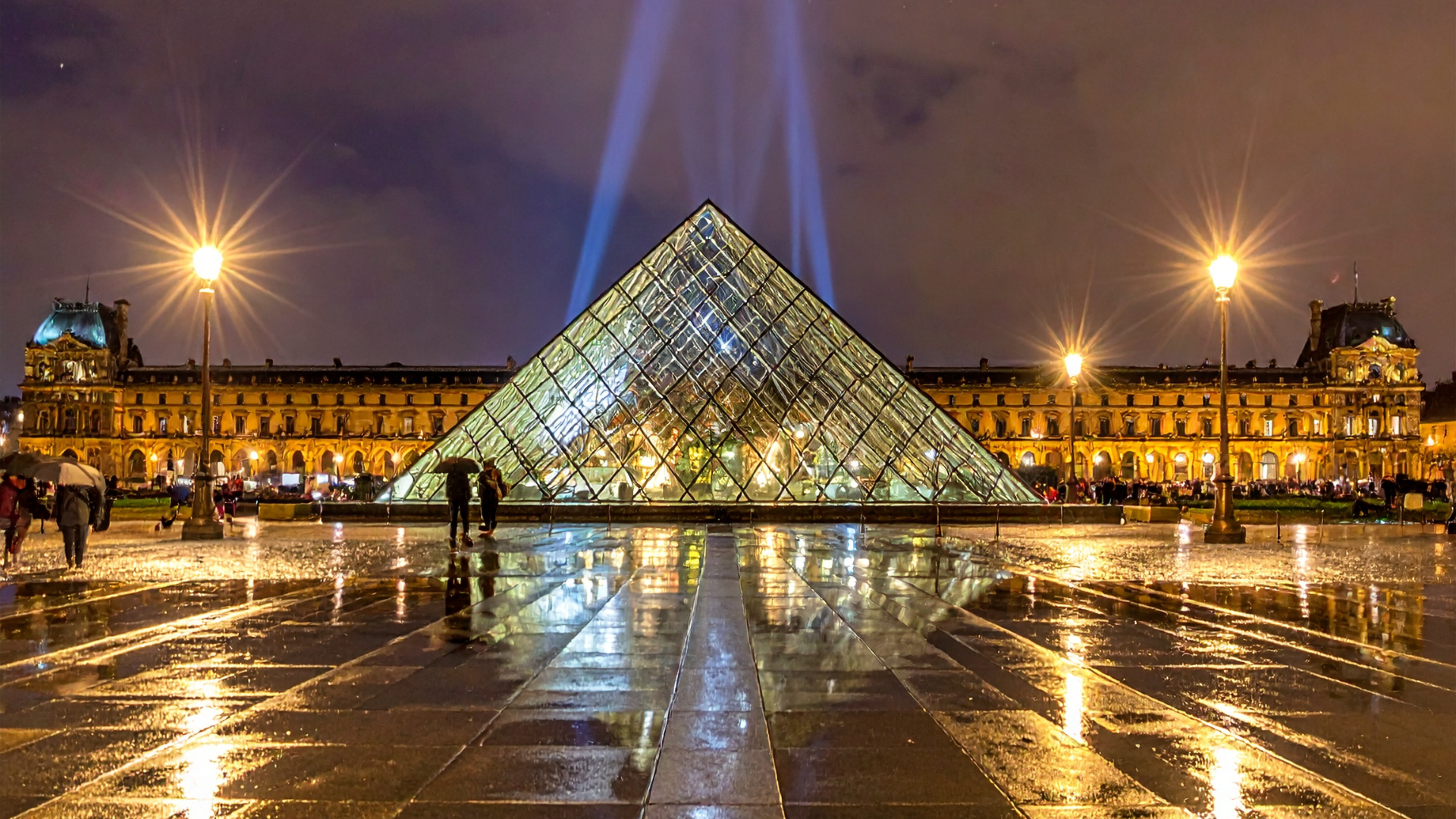 Illuminated glass pyramid of the Louvre Museum at night, reflecting on rain-soaked paving stones, with historic buildings and glowing street lamps in the background