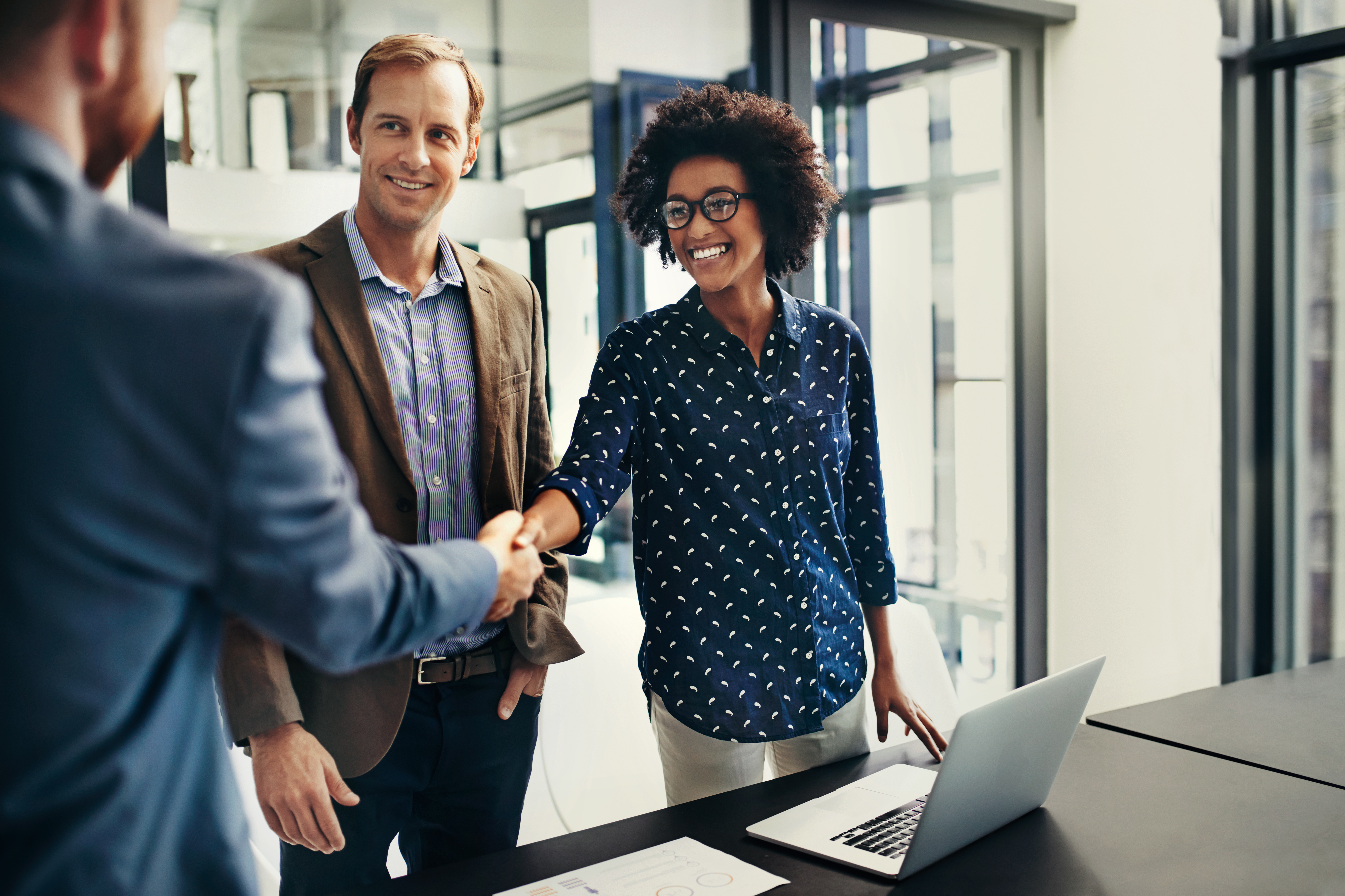 Image of man, woman and a figure partially visible, reaching his hand forward to handshake the woman. They are wearing with business clothes and have a laptop on the desk in front of them. 